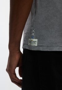 Γκρι βαμβακερό μπλουζάκι με πλαϊνή ετικέτα και κεντημένες λεπτομέρειες. Διαθέτει άνετη εφαρμογή και εμφανή ραφή στο τελείωμα. Μερικώς εμφανισμένο μαύρο παντελόνι.