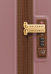 Close-up van een roze koffer rits met gouden SUITSUIT-tags en een combinatieslot, met zichtbare bruine stiksels naast de rits.