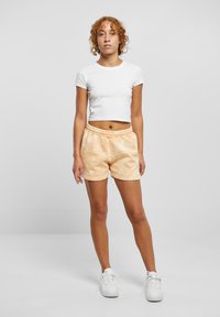Hvit avkortet t-skjorte kombinert med ferskenfargede tie-dye shorts med elastisk midjebånd. Modellen med hvite joggesko står mot en ensfarget bakgrunn.