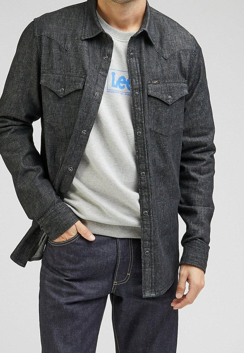 Camisa de mezclilla oscura con botón en la parte delantera, dos bolsillos en el pecho y hombros de estilo vaquero, superpuesta sobre una sudadera gris claro.
