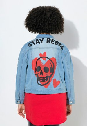 Person med krøllet hår iført lyseblå denimjakke med teksten "STAY REBEL" og rødt kranie- og hjerte-design på ryggen, rød kjole nedenunder.