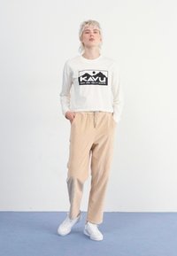 Weißes Langarmhemd mit schwarzem KAVU-Logo, kombiniert mit gestreiften beigen Hosen. Das Outifit wird mit weißen Turnschuhen abgerundet. Einfaches Design.