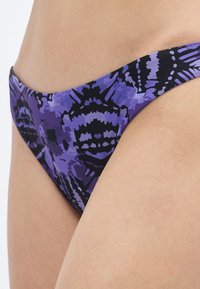 Μπικίνι κάτω σε μοτίβο μωβ και μαύρου με ομαλή υφή, μεσαία μέση και μινιμαλιστική κοπή. Διαθέτει καλλιτεχνικές γραφικές λεπτομέρειες.