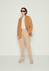 Brun kavaj över en beigefärgad topp, kombinerad med ljusbeige höga byxor och tan plattformssandaler. Fyrkantiga svarta solglasögon fulländar looken.