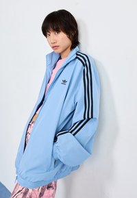 Chaqueta azul claro con cremallera y diseño de tres rayas negras en las mangas, puños acanalados y un pequeño logo negro en el pecho. Textura suave.