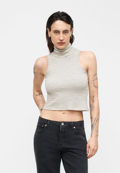 Personne aux cheveux courts et foncés, portant des boucles d'oreilles, un haut court à col roulé rayé sans manches et un jean noir, montrant des tatouages sur les deux bras.