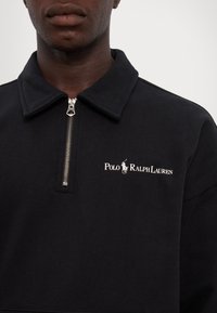 Svart tröja med silverfärgad dragkedja i kragen, med en vit "Polo Ralph Lauren"-logotyp. Tillverkad av mjukt material, med en enkel design och avslappnad passform.