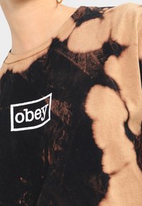 Short-sleeve, tie-dye shirt with brown and black patterns, featuring a white rectangular logo reading "obey" on the front. Soft fabric texture.