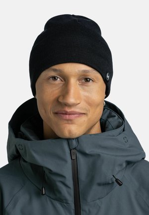 Black knitted beanie with a ribbed texture and a small logo tag, worn with a dark green zip-up jacket featuring a hood.
