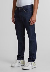 Jeans de mezclilla oscura con un corte recto, diseño de cinco bolsillos y costuras doradas visibles. Combinados con zapatillas blancas y grises.