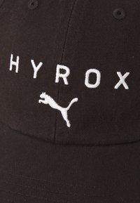 Sort kasket lavet af bomulds-twill, med hvid broderet tekst "HYROX" og et springende Puma-logo på fronten. Glat tekstur, buet skygge.