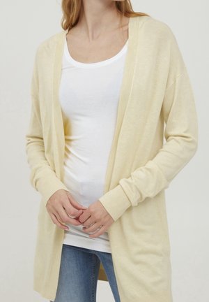 Jasnożółty kardigan zapinany z przodu, z ribbingu na mankietach, o miękkiej fakturze i długich rękawach, noszony na białej dopasowanej koszuli i niebieskich dżinsach.