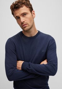 Navy blue knit sweater with crew neckline, ribbed cuffs, and a small tonal brand logo on the chest; smooth texture, fitted design.