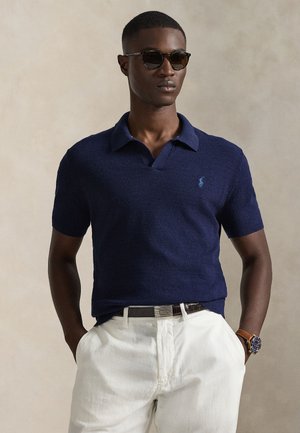 Μπλε πλεκτό πόλο μπλουζάκι με γιακά και κοντά μανίκια, με μικρό μπλε λογότυπο στο στήθος. Συνδυασμένο με άσπρο παντελόνι.