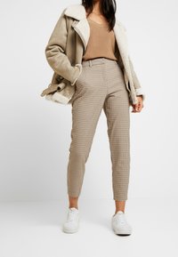 Beżowa kurtka pilotka wyłożona beżowym kożuszkiem, noszona na brązowym prążkowanym topie, zestawiona z wysokim stanem spodni w pepitkę i białymi trampkami.