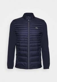 Marinblå jacka med vadderad framsida och släta ärmar. Har dragkedjestängning, ståkrage och liten grön logotyp på vänster bröst.