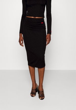 Woman wearing a black pencil skirt and long-sleeve crop top, paired with black strappy heeled sandals, standing against a plain white background.