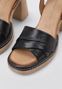 Sandalias de tacón de cuero negro con correas entrelazadas, plantillas en tono marrón y un tacón grueso de color claro. Presenta costuras blancas contrastantes.