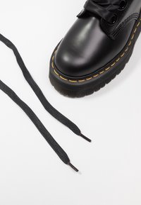 Bota de cuero negro con punta redonda, cordones planos negros, suela gruesa negra y costuras amarillas alrededor de la suela. Dos cordones sueltos al lado de la bota.