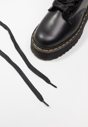 Botte en cuir noir à bout rond, lacets plats noirs, semelle épaisse noire et coutures jaunes autour de la semelle. Deux lacets lâches à côté de la botte.