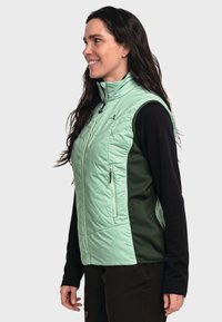 Frau lächelt und trägt eine hellgrüne gesteppte Weste mit Reißverschlusstaschen über einem schwarzen Langarmshirt, während sie vor einem weißen Hintergrund steht.