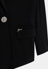 Svart kavaj i slätt tyg, med en silverknapp, strukturerade axlar och en ficka med "Guess"-logotyp.
