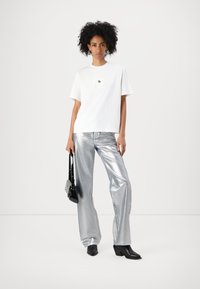 Weißes Baumwoll-T-Shirt mit einem perforierten quadratischen Design, kombiniert mit elfenbeinfarbenen weiten Hosen. Schwarze Absatzstiefel und kleine schwarze Tasche.