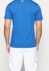 Muž v jednoduchém modrém tričku s krátkým rukávem a oranžovým logem Under Armour na horní části zad, s bílými kalhotami s černými logy.