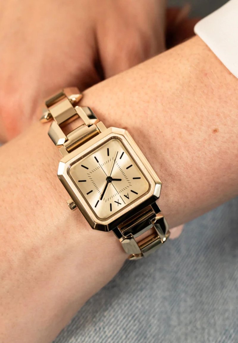 Goldene quadratische Armbanduhr mit römischen Ziffern und schwarzen Zeigern, getragen am linken Handgelenk mit heller Haut vor einem blauen Stoffhintergrund.