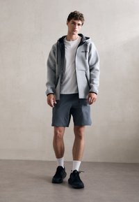 Siva zip-up dukserica s crnim detaljima preko bijele majice, u kombinaciji sa sivim šortsevima i crnim tenisicama. Postavljena na neutralnoj pozadini.