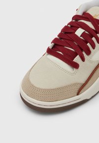 Bézs sportcipő velúr felsőrésszel, piros fűzőkkel és perforált orr résszel. Figyelemre méltó részletek: barna gumitalp és narancssárga díszítések.