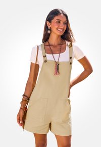 Beige tuinbroek shorts van een katoenmix, met grote voorzakken, verstelbare banden en knoopaccenten. Draagt over een witte top.