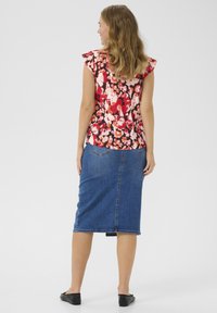 Blouse florale en rouge, rose et blanc avec des manches volantées ; associée à une jupe crayon en jean bleu. Chaussures plates noires visibles.