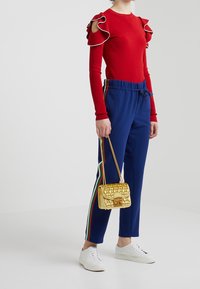 Maglietta rossa a maniche lunghe con spalle arricciate, pantaloni blu con strisce laterali multicolori e una piccola borsa a tracolla trapuntata dorata. Sneakers bianche.
