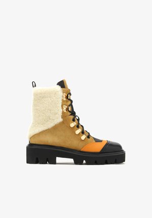 Lace-up ankle boots featuring a tan suede upper with black and orange accents, plush shearling collar, chunky black rubber sole, and gold hardware.
