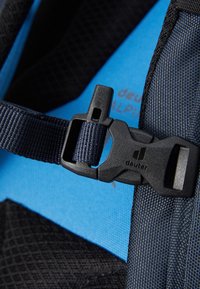 Black and blue backpack strap with a plastic buckle, featuring woven texture and a logo. Blue interior visible in the background.