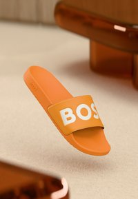 Πορτοκαλί σαγιονάρα με πλατιά λουρά, διακοσμημένη με τη λέξη "BOSS" σε λευκό. Κατασκευασμένη από μαλακό υλικό με υφή για καλύτερο κράτημα.