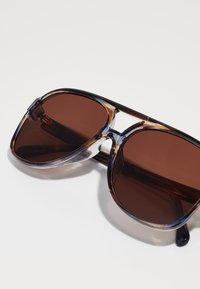 Ochelari de soare cu un cadru bicolor, având lentile de culoare maro închis și un model transparent de scoică țestoasă, evidențiind o formă modernă rectangulară.