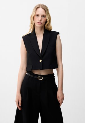 Donna con capelli biondi lunghi che indossa un blazer nero corto senza maniche con un bottone dorato e pantaloni neri a vita alta con cintura.