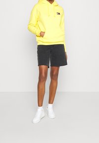 Felpa gialla con tasca frontale e cordino, abbinata a pantaloni corti in denim neri. Sneakers bianche completano il look.