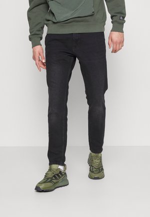 Personne portant un jean noir slim, des baskets vertes et noires, et un haut vert à manches longues, debout devant un fond uni.