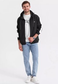 Felpa nera con zip e dettagli bianchi, indossata sopra una t-shirt bianca con una grafica. Jeans azzurri chiari e sneakers bianche completano l'outfit.