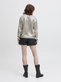 Λαμπερό ασημί πουλόβερ με μακριά μανίκια, υφασμένο με υφή, συνδυασμένο με σκούρες βερμούδες και μαύρες δερμάτινες μπότες μέχρι το γόνατο σε ουδέτερο φόντο.