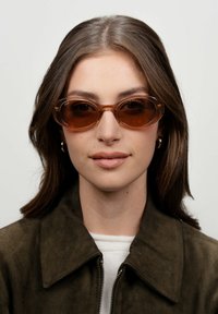 Gafas de sol ovaladas con un tinte marrón y montura transparente. Las monturas tienen curvas sutiles y un acabado brillante. Se llevan con una chaqueta de ante verde oliva.