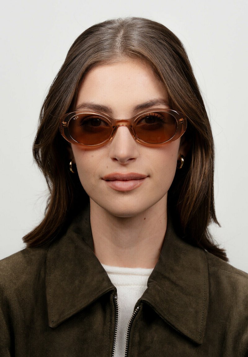 Gafas de sol ovaladas con un tinte marrón y montura transparente. Las monturas tienen curvas sutiles y un acabado brillante. Se llevan con una chaqueta de ante verde oliva.
