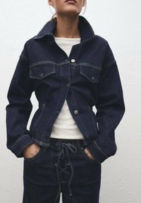 Tamni traper jakna s dva džepa na grudima, zatvaranjem na dugmad i vidljivim šavovima. Nosit će se preko bijele majice, u kombinaciji s tamnim traper hlačama.