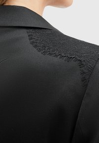 Gros plan sur l'épaule d'une veste noire avec des détails en dentelle le long de la couture et du col, portée par une personne de dos.