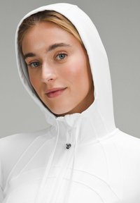 Haut de sweatshirt zippé blanc avec col montant et cordons de serrage réglables. Présente un tissu lisse et texturé avec un détail de logo subtil.