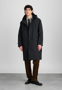 Black waterproof coat with a hood, featuring two front pockets and a button closure. The model wears brown corduroy pants and black shoes.