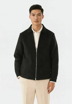 Svart, texturerad zip-försluten jacka med krage, med sidofickor och en subtil glans. Bärs över en vit, krage skjorta.